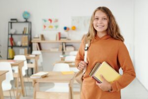A cheerful schoolgirl in an orange jacket stands in a bright classroom holding notebooks, ready for a new school year.
